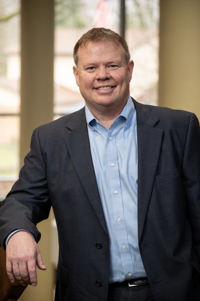 A middle-aged man in a casual suit and button-up shirt leaning on a banister.